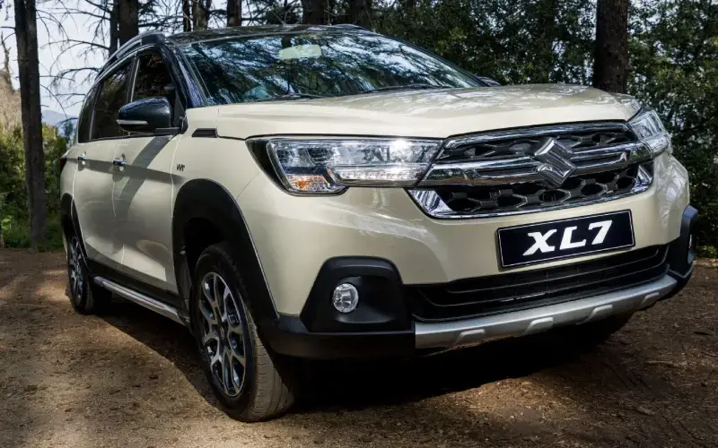 Suzuki XL7, acabado exterior en color beige, mostrando su parrilla frontal y faros en un paisaje natural.